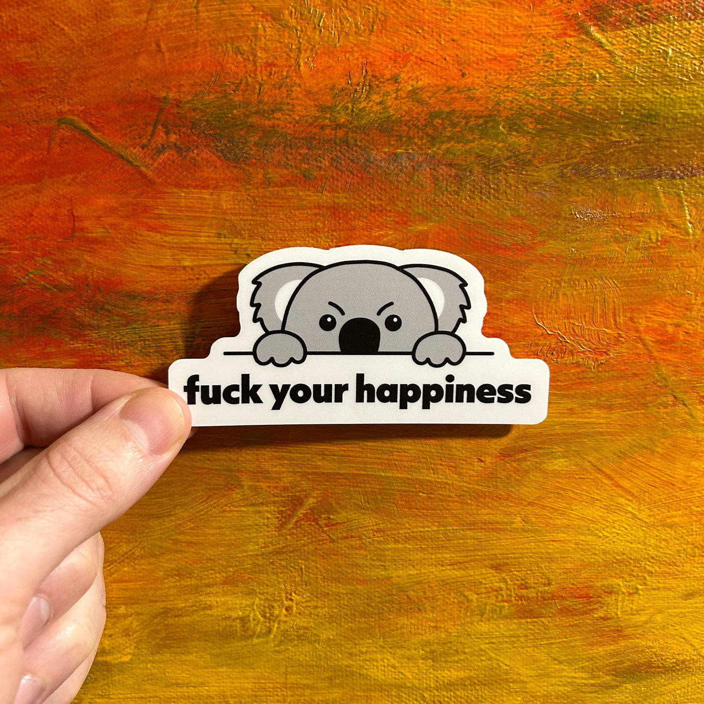 Sticker of an illustrative koala head with a cute kawaii angry face lifting its head over a ledge so you can’t see its mouth. Just under the ledge it says “fuck your happiness”.