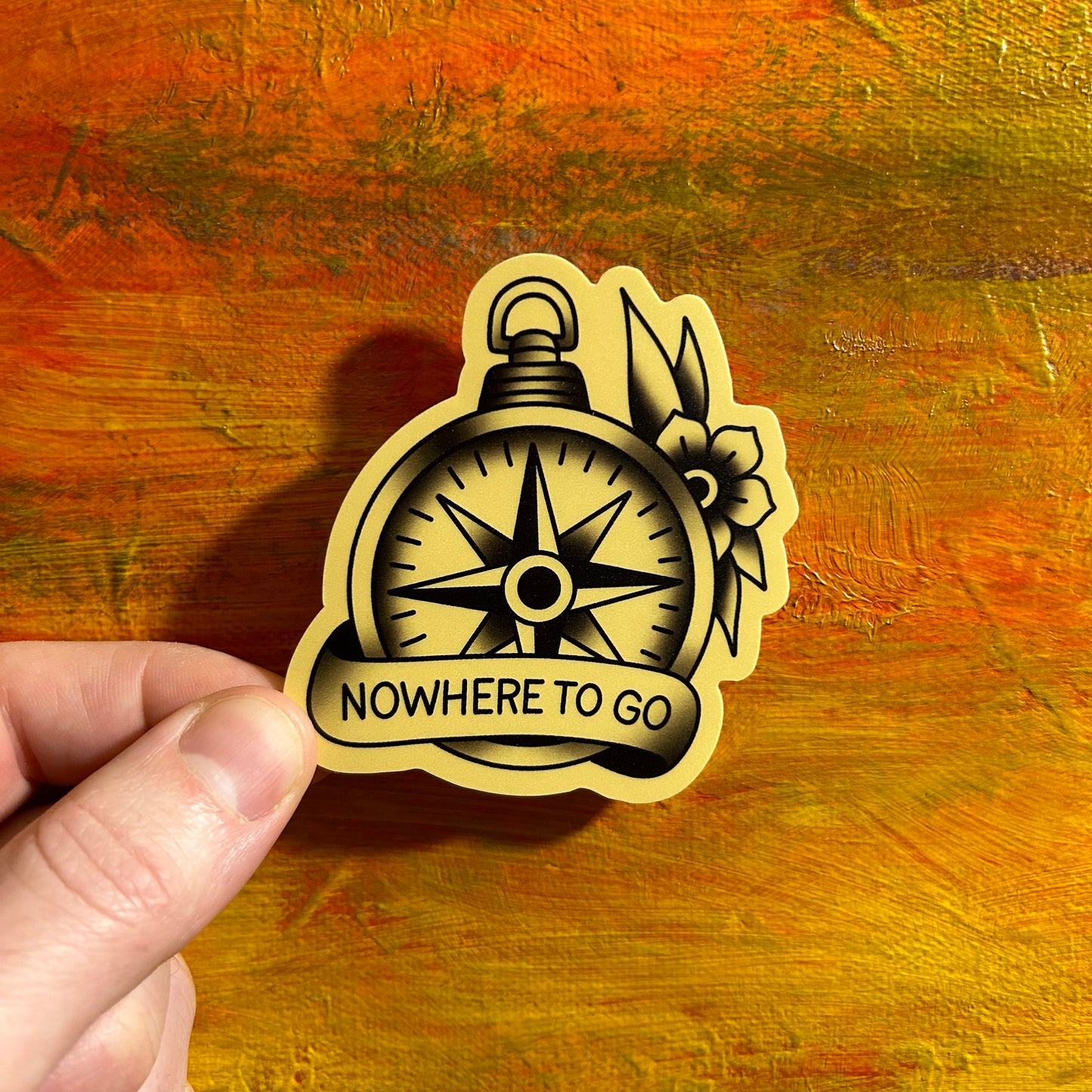 Tattoo style die cut sticker drawing of a black and grey compass with a banner across the bottom part saying “NOWHERE TO GO”. There is a black and grey flower coming out from behind the compass in the top right corner.