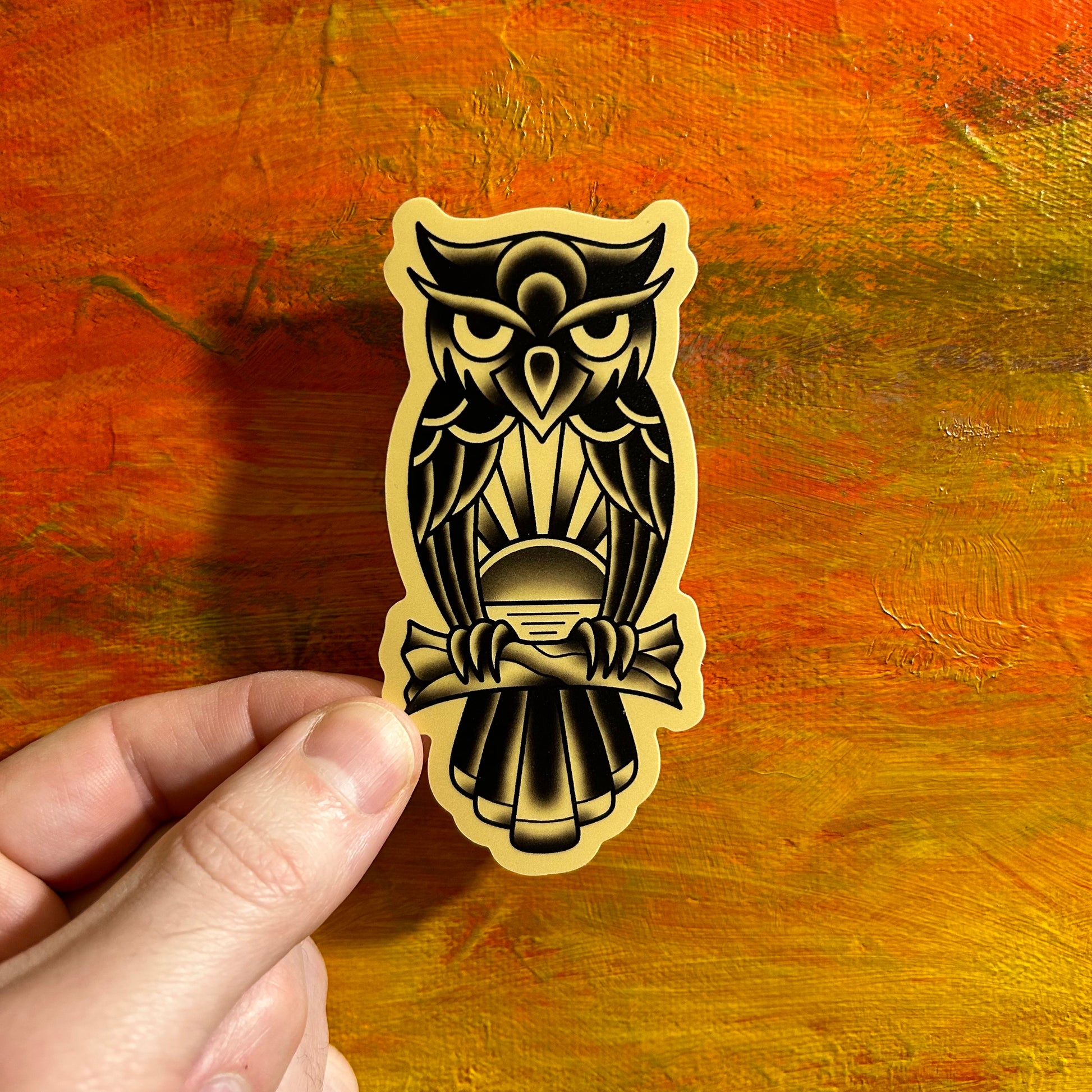 A die cut sticker of a black and grey owl with a sunset theme in its chest. The owl is standing on a branch and the design is high contrast, bold and tattoo style.