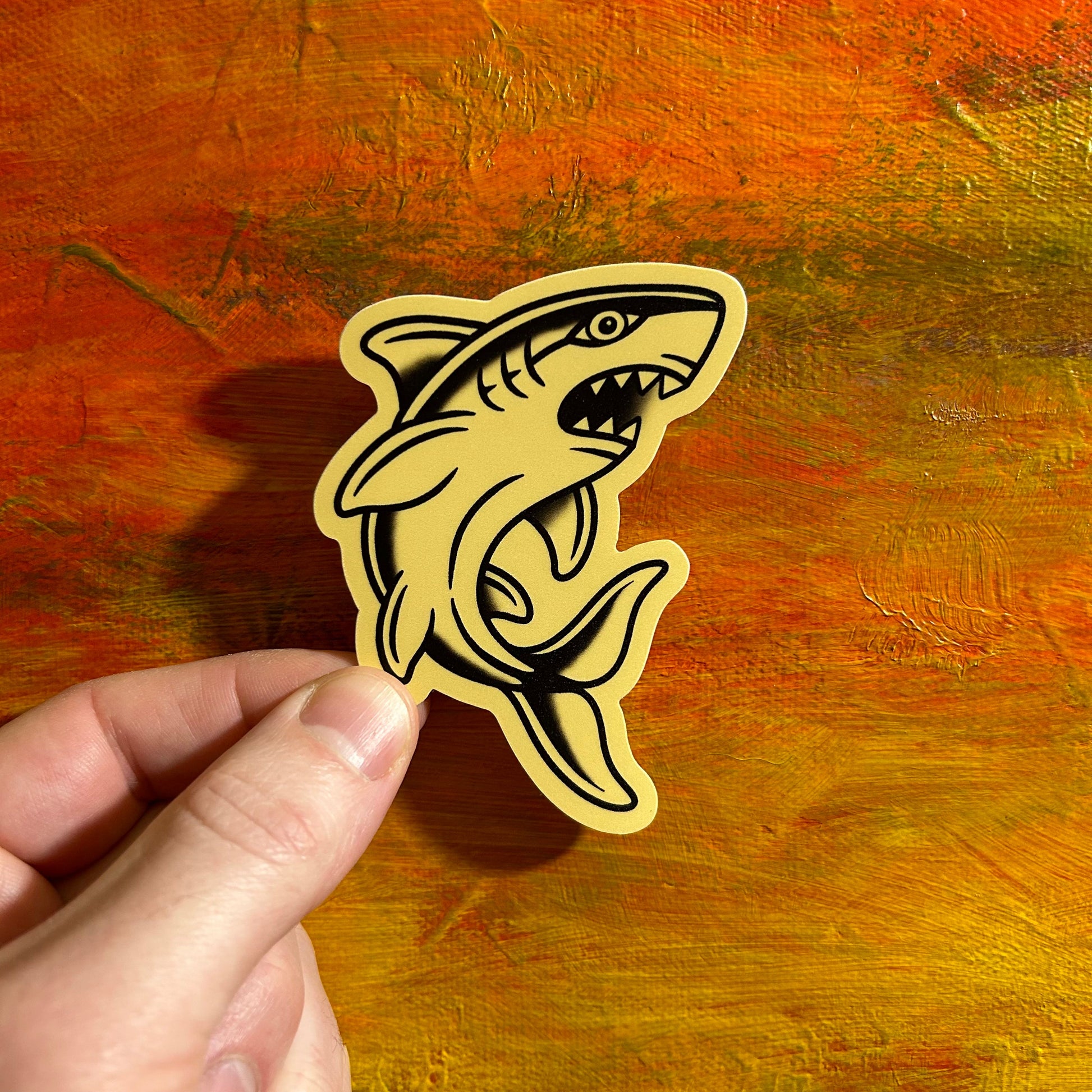 Sticker of an illustrative shark on tan background. The sticker is die cut to the shape of the shark.