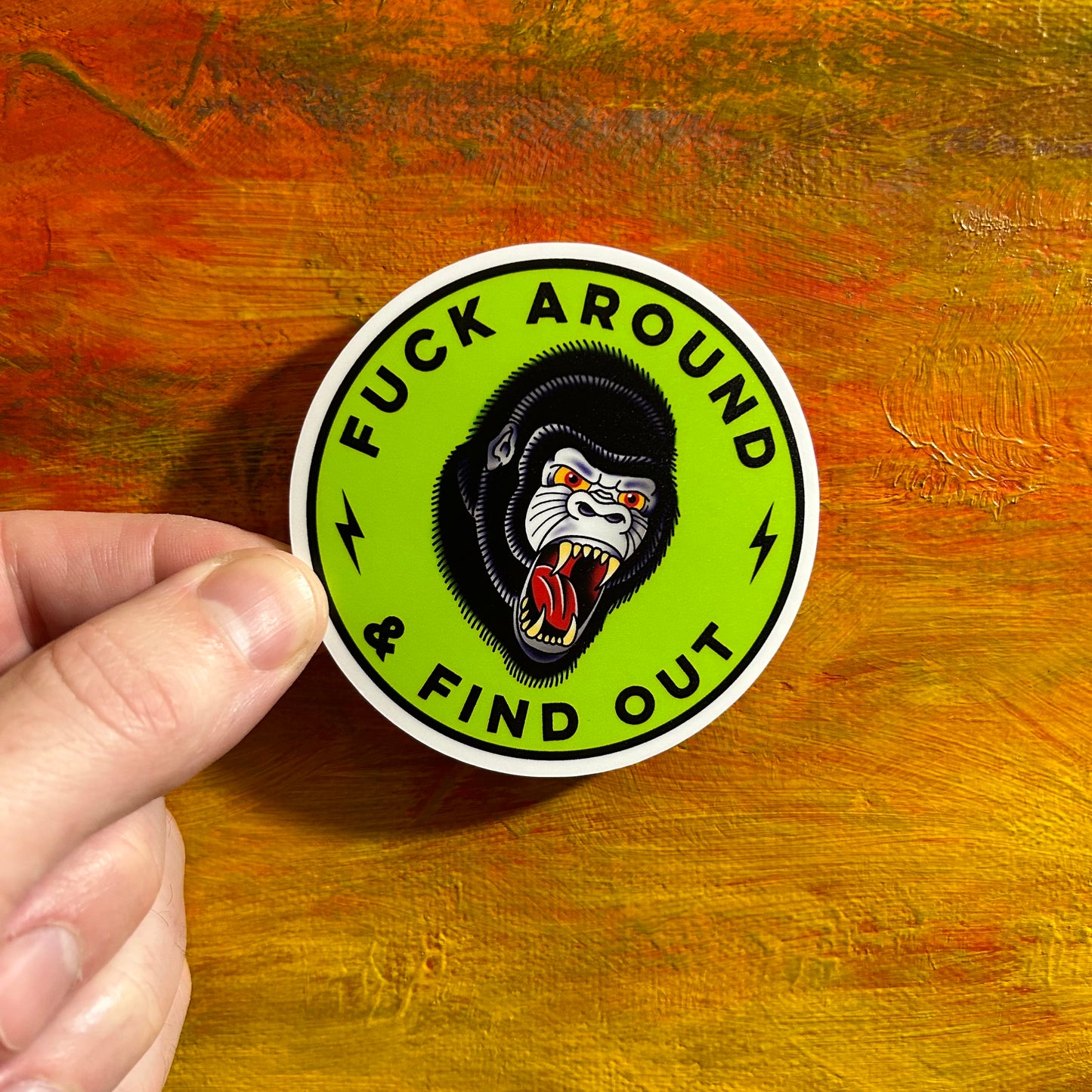 Round bright green sticker with a drawing of a black gorilla head with its menacing open mouth look and piercing eyes. The text wrapped around the gorilla says “FUCK AROUND & FIND OUT”.