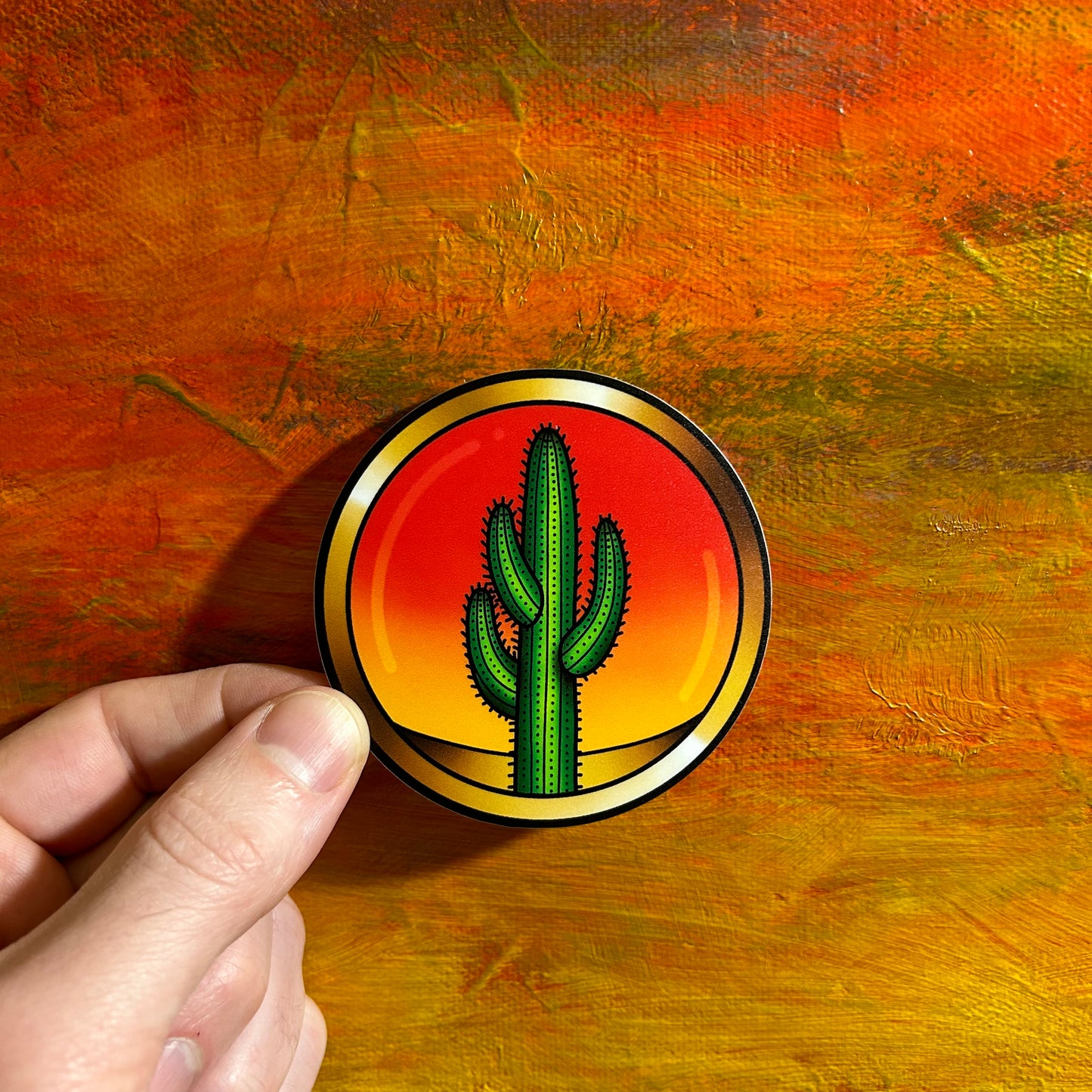 A round sticker of yellow and red gradient sky with an illustrative green cactus in the foreground. The drawing gives the illusion that the scene is set in a bubble frame.