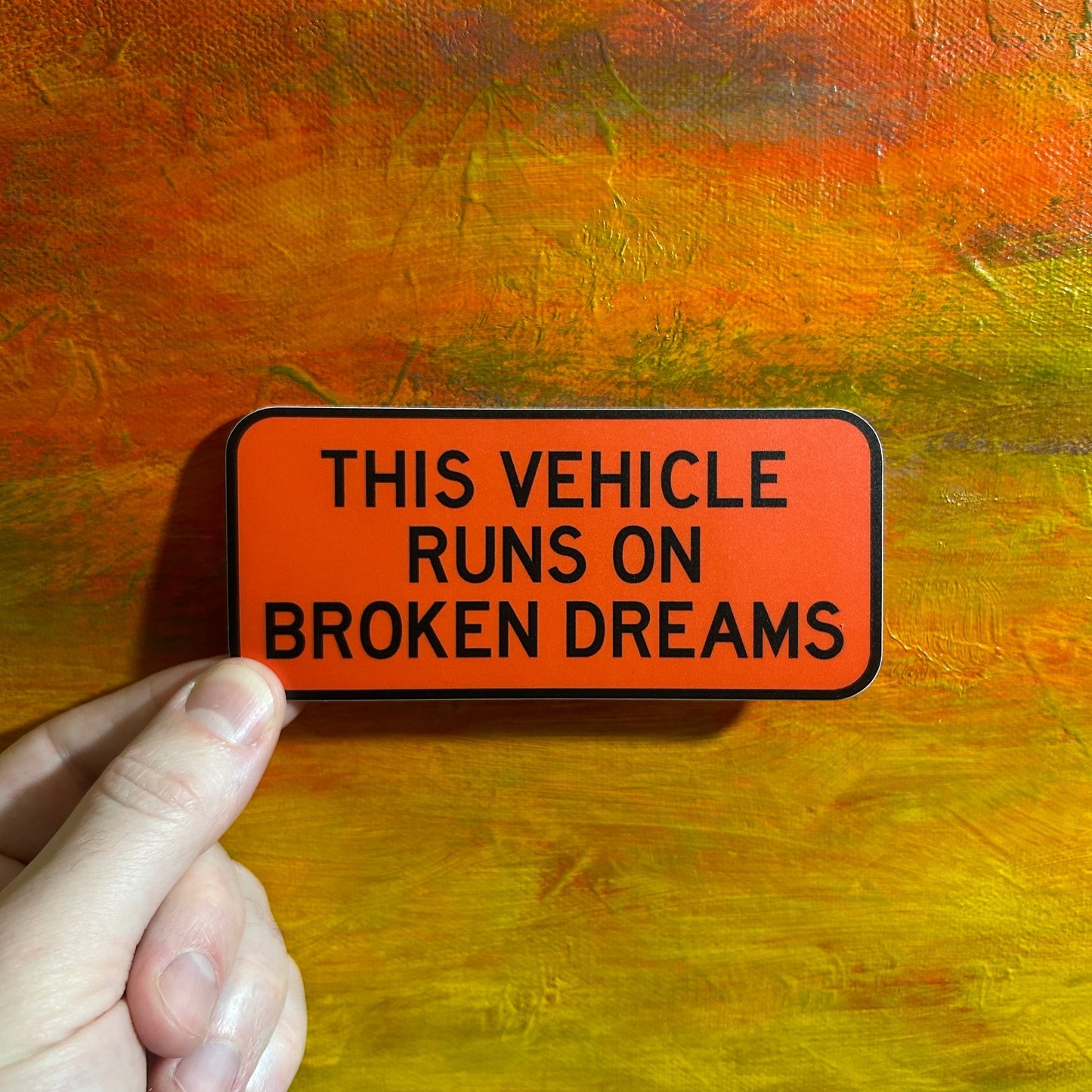 Rectangular bright orange sticker with black construction lettering saying “this vehicle runs on broken dreams”.