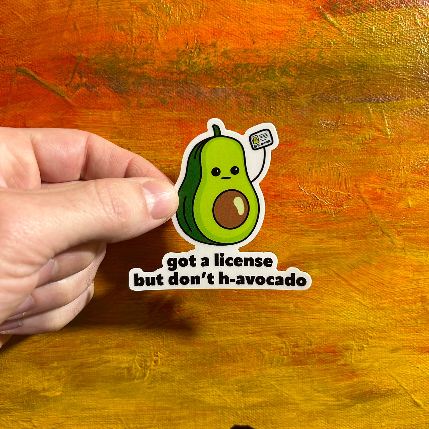 Sticker of a comical avocado with a cute mellow kawaii face holding up a drivers license. Underneath it is “got a license but don’t have h-avocado”.