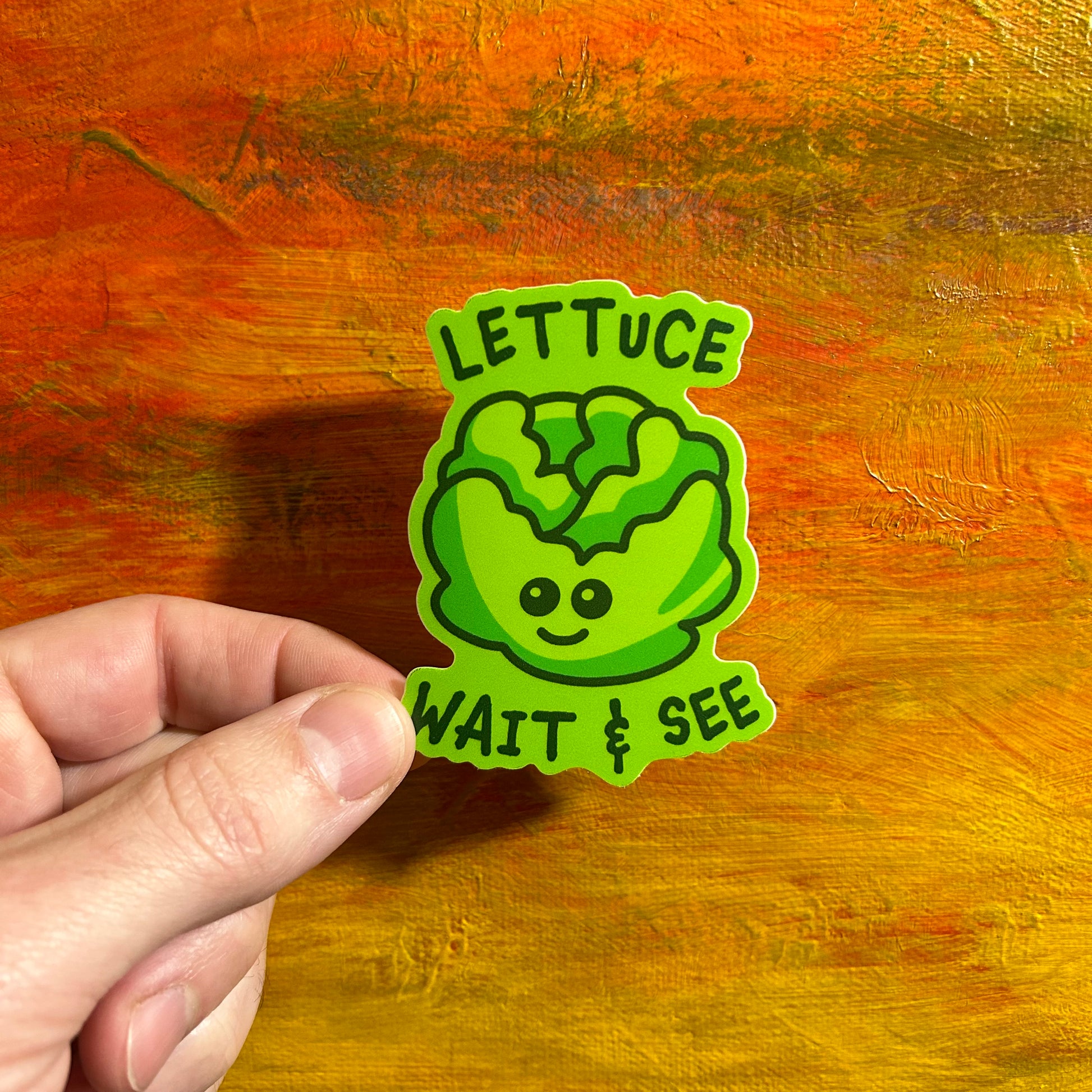 Illustrative comical sticker of a funny green head of lettuce with a little kawaii cute smiley face. The lettuce is adorned with the saying “lettuce wait & see”.