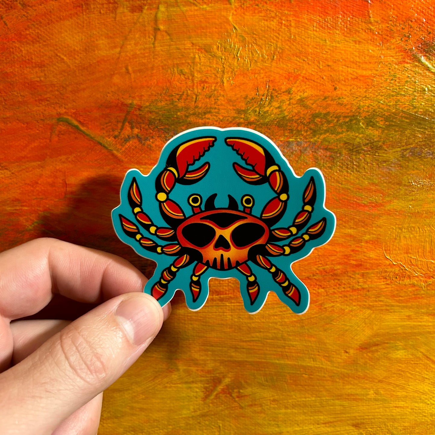 Sticker of a red yellow crab in an illustrative tribal style with the markings of a skull on the crabs back. The background around the crab is a dark teal.