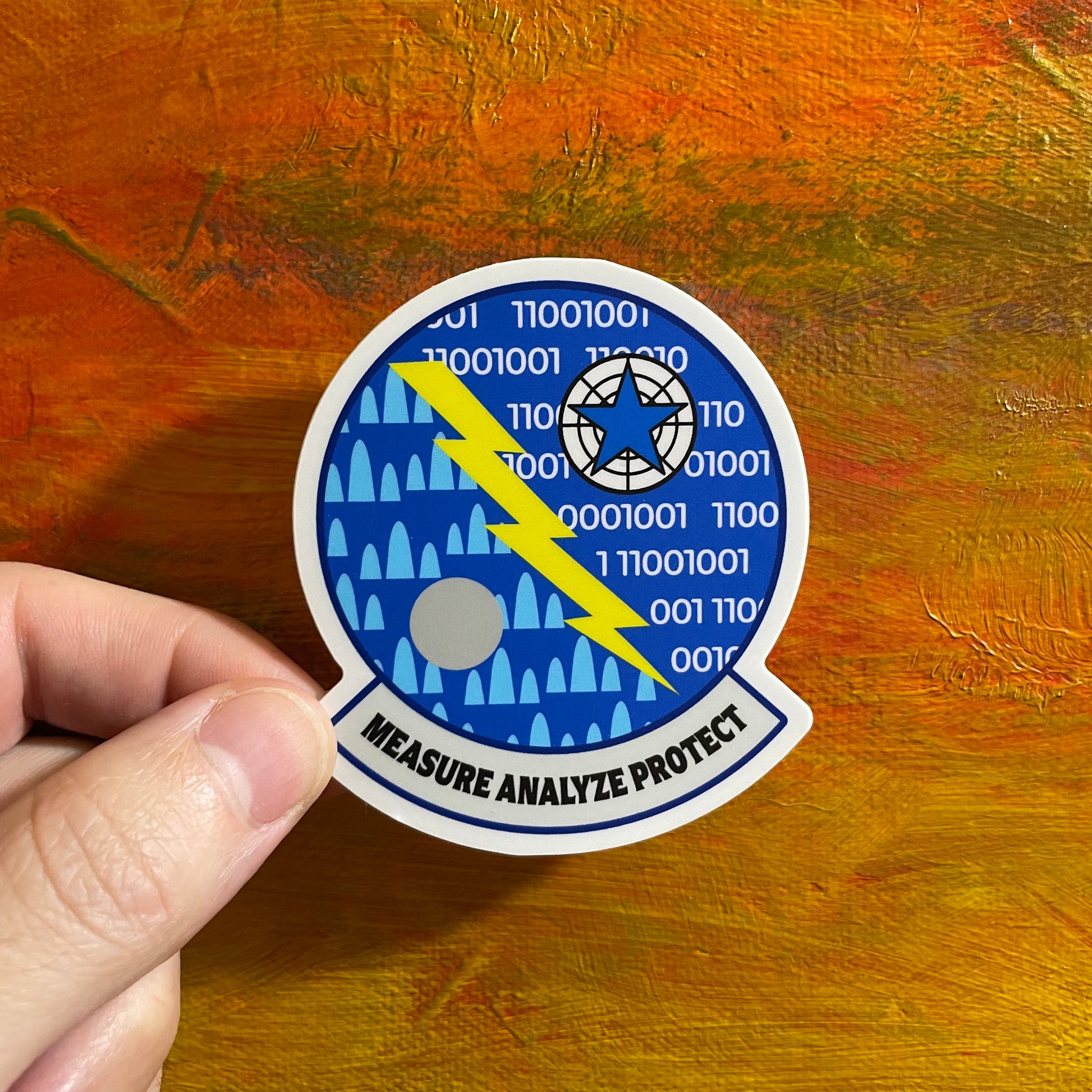 A sticker of a military type logo patch. Blue background with a yellow lightning bolt that divides two sides of the logo. A lot of intricacies with binary code and shapes that form an ambiguous design shrouded in mystery. At the bottom it says measure analyze protect.