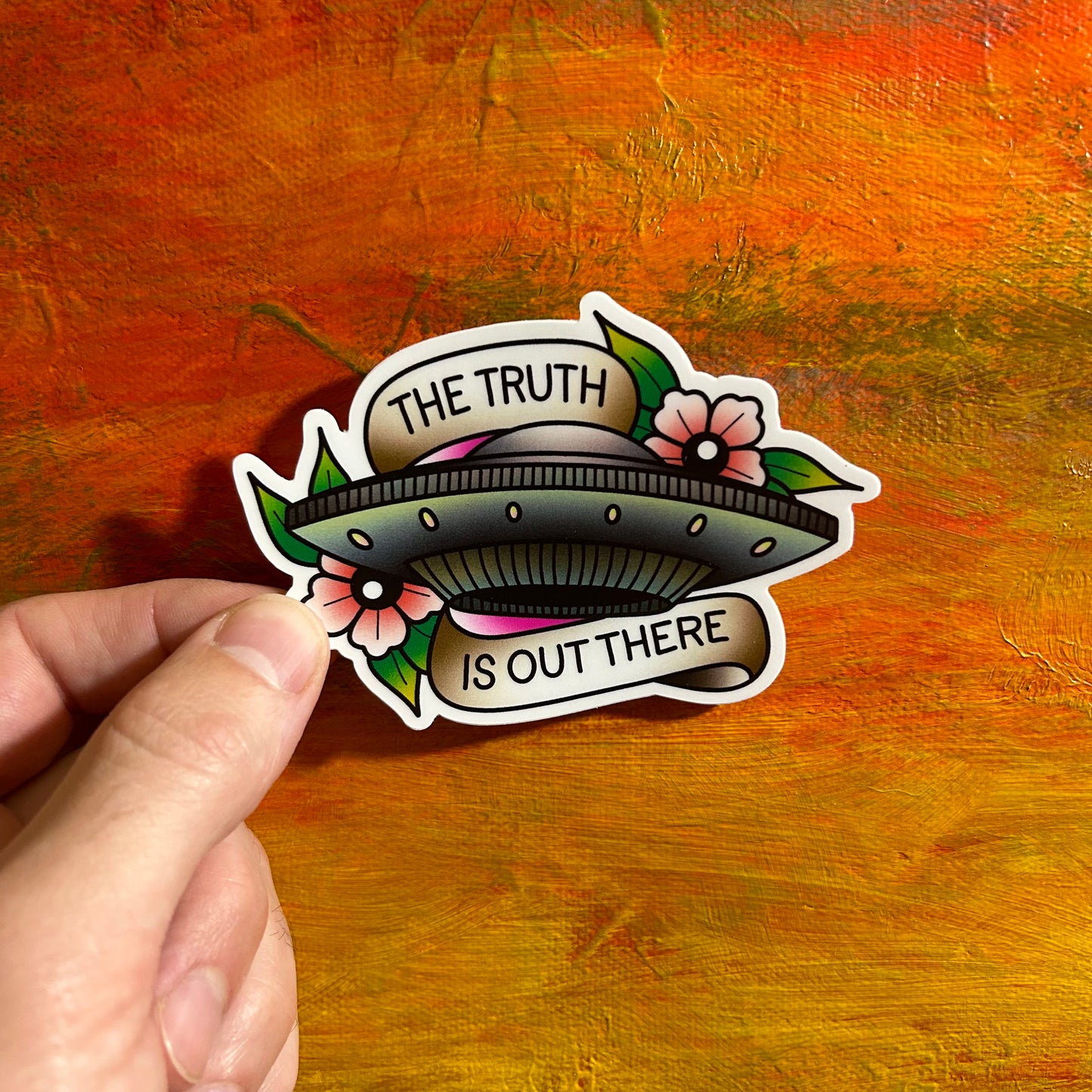 A ufo alien spaceship adorned by colourful water lillies on both sides, an old school tattoo banner wrapped above and below saying “the truth is out there”