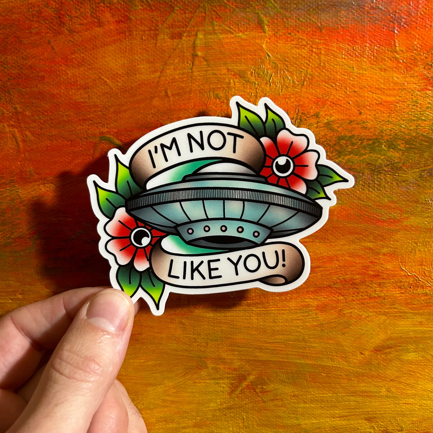 The image shows a hand holding a sticker of a UFO with a banner reading "I'M NOT LIKE YOU!" The design is framed by flowers and leaves in a tattoo-like style. An ode to Blink 182 and my love for UFOs