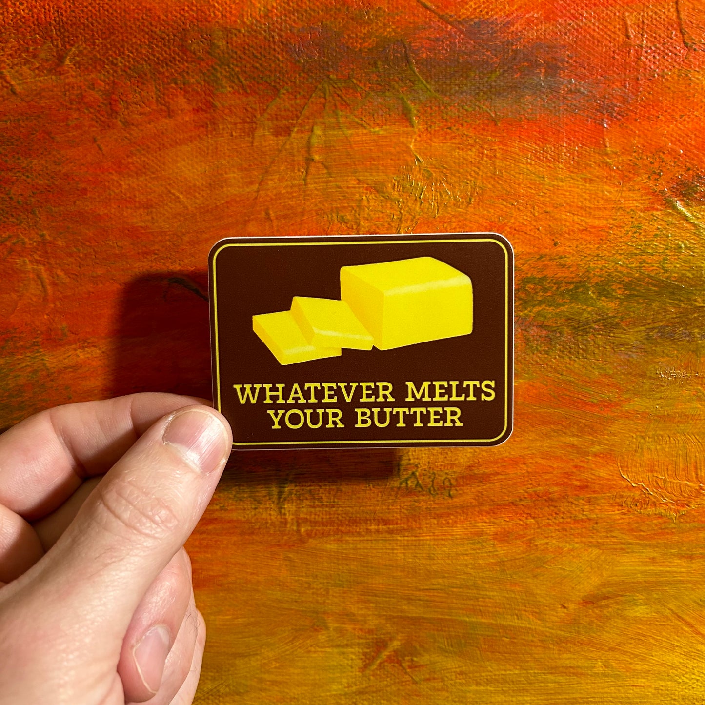 A dark brown rectangular sticker with an illustration of soft yellow butter and below it says “whatever melts your butter”.