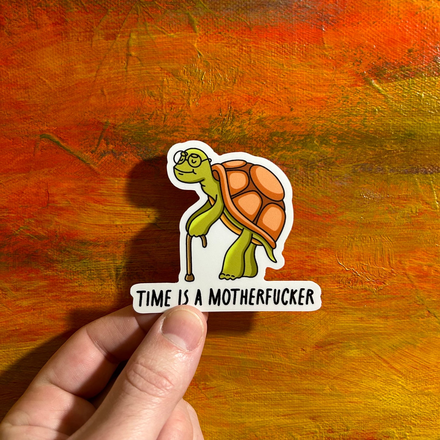 Die cut sticker of an illustrative old turtle with a cute smile. Underneath the turtle it says “time is a motherfucker”.