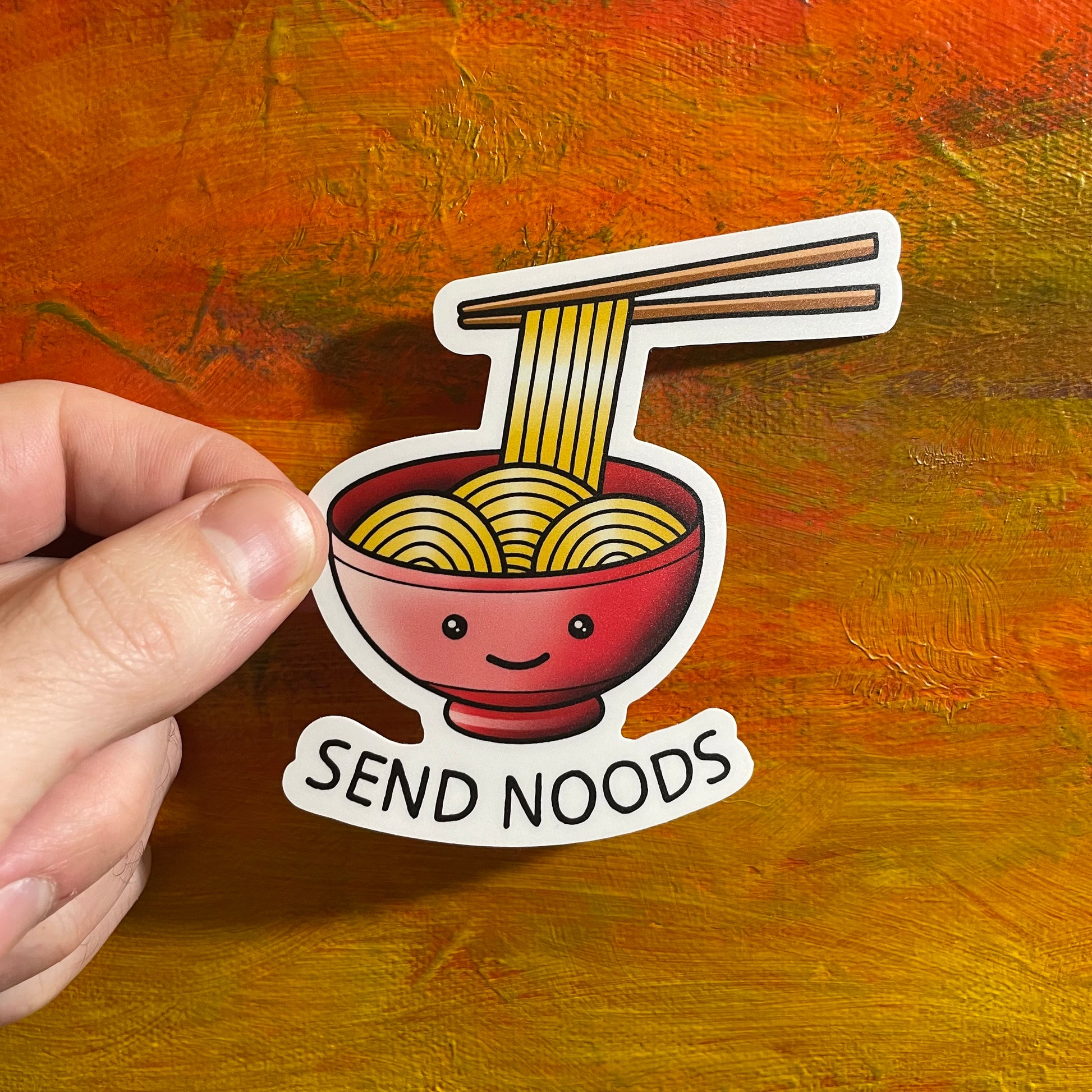 A die cut sticker of an illustrative drawing of a bowl of noodles with a cute face. There is a set of chopsticks lifting up some noodles and underneath the red bowl with the smile says “send noods”.