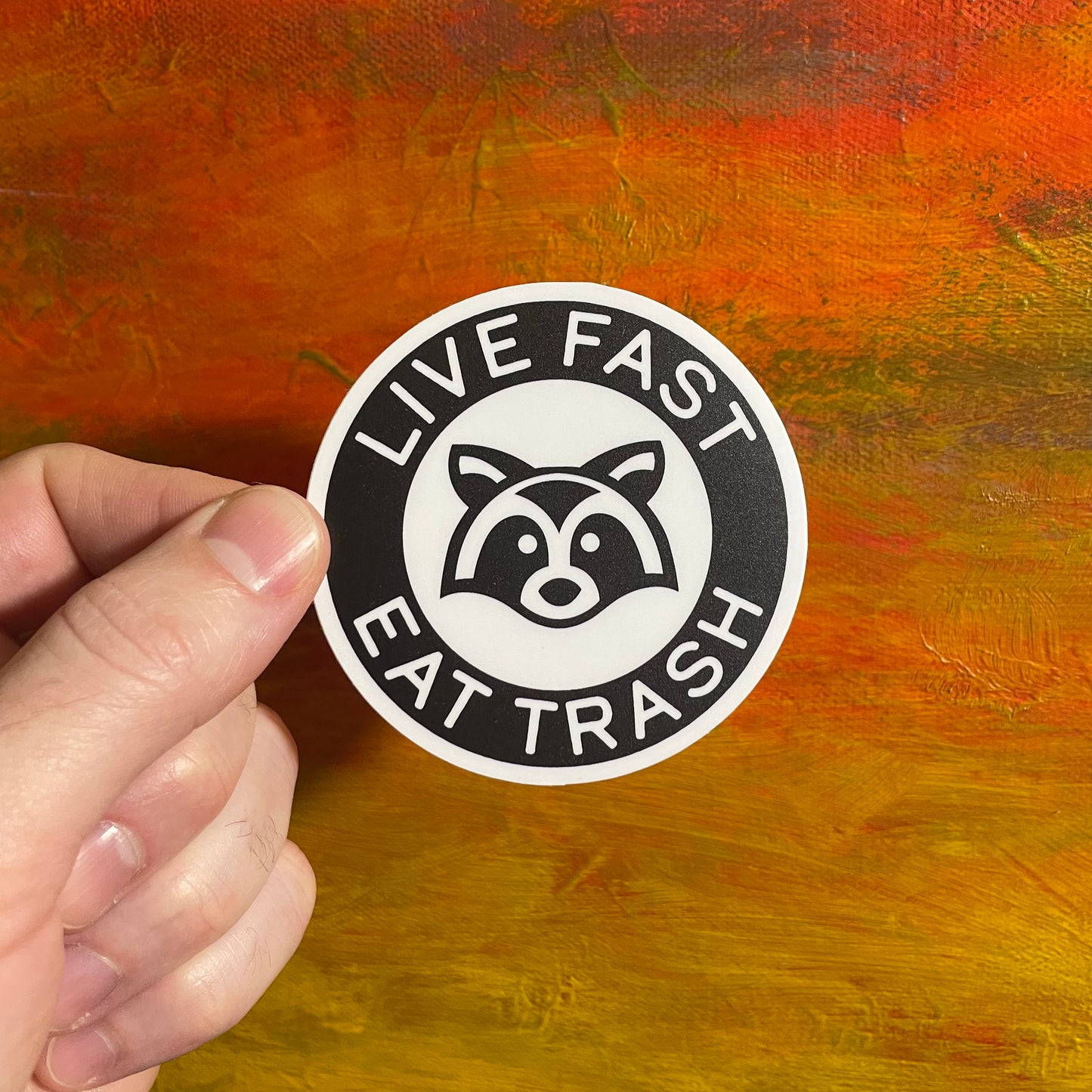 Bold contrast black and white Illustration of a raccoon face. Font is wrapped around the top and bottom in arches saying “live fast eat trash”. Circle sticker.