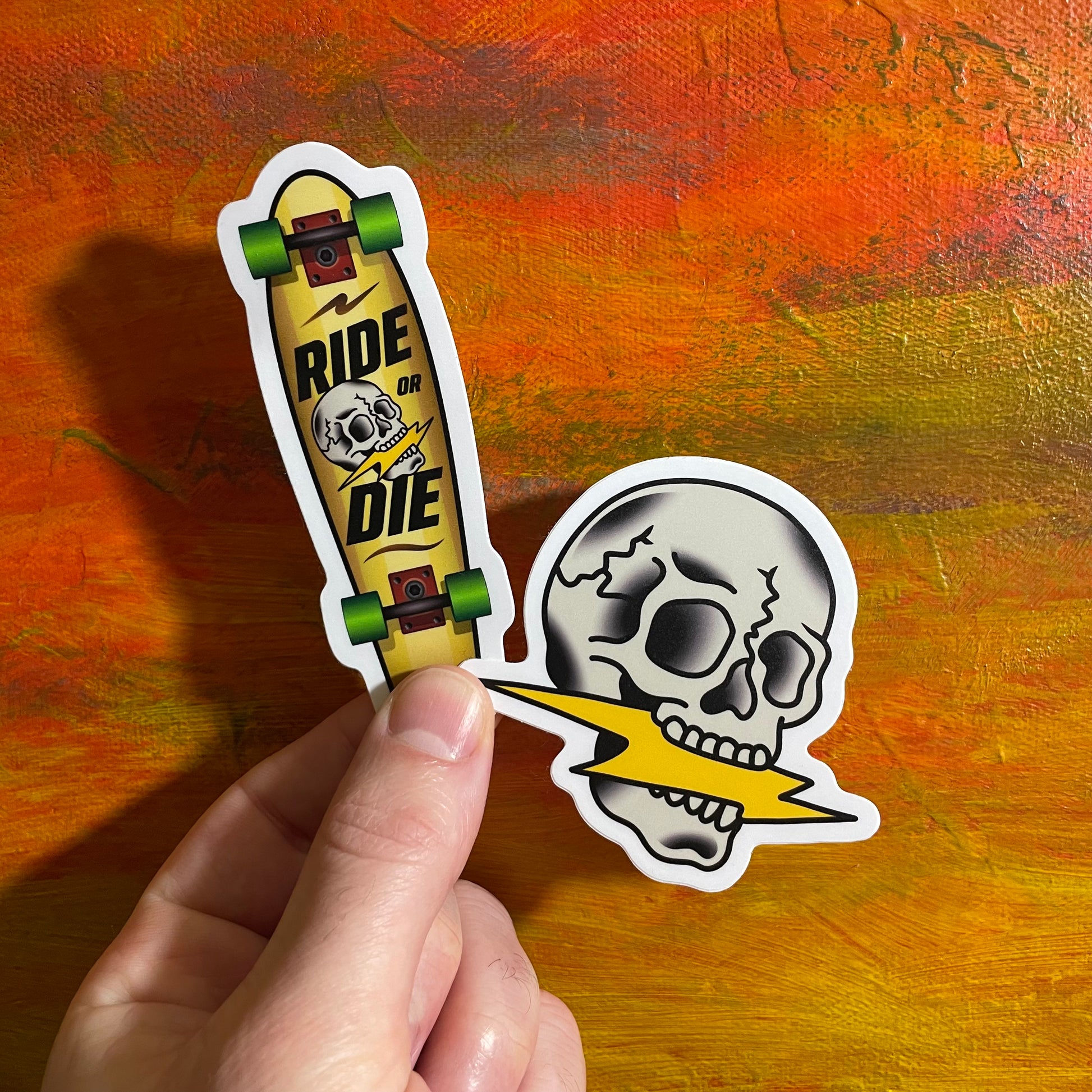 Sticker of a grey skull biting on a yellow lightning bolt. The sticker next to it is a yellow tan skateboard with green wheels and red trucks. On the deck it has the skull eating lightning design and says RIDE OR DIE.