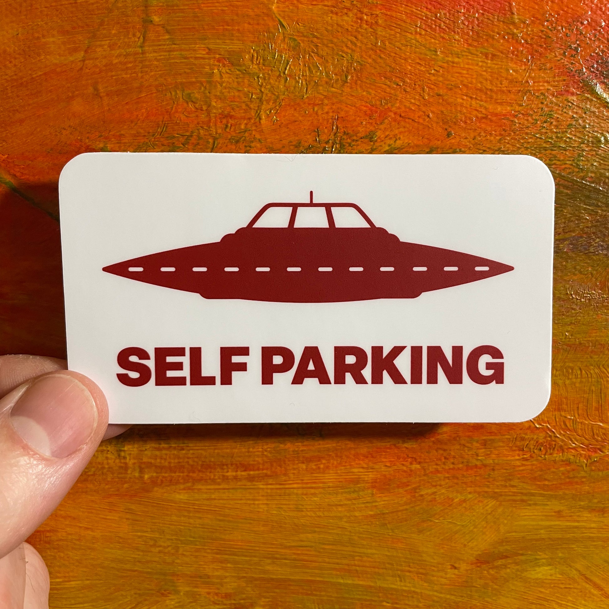 A white rectangular waterproof sticker with a red UFO spacecraft and red capital letters underneath saying “self parking”