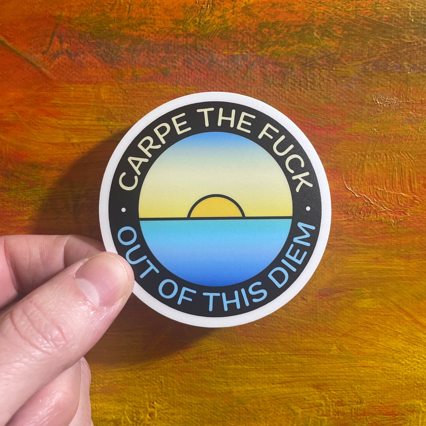 A circle sticker of a minimal illustrative sunset over a lake and a saying arched over the top half and bottom half saying “carpe the fuck out of this diem”.