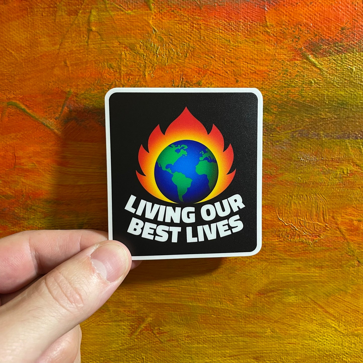 A square black sticker with an illustration of earth burning and below it says “living our best lives”.